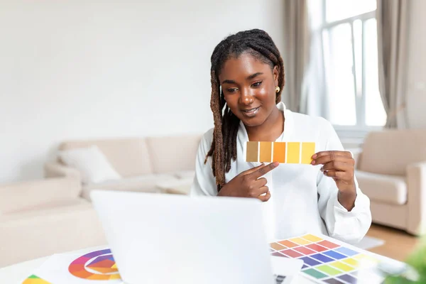 Cheerful african american lady designer having video conference with clients, sitting at desk in front of computer, holding color palettes, gesturing and smiling, copy space