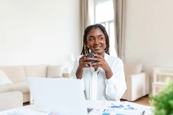 Young African American businesswoman working remotely from home using a laptop and enjoying a cup of coffee, smiling