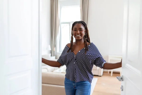 Welcome. Portrait of cheerful African woman inviting visitor to enter his home, happy young woman standing in doorway of modern apartment showing living room with hand