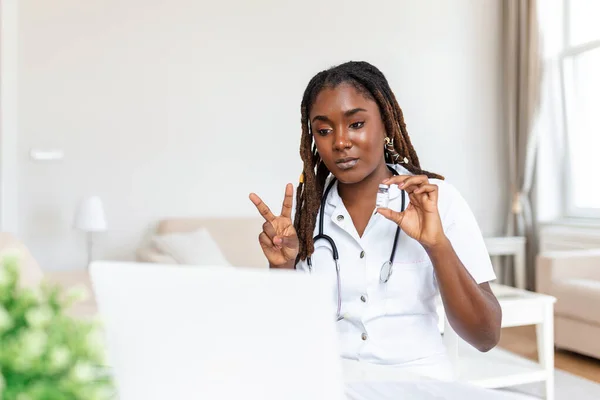 African woman doctor talking online with patient, making video call, looking at camera, young female wearing white uniform with stethoscope speaking, consulting and therapy concept