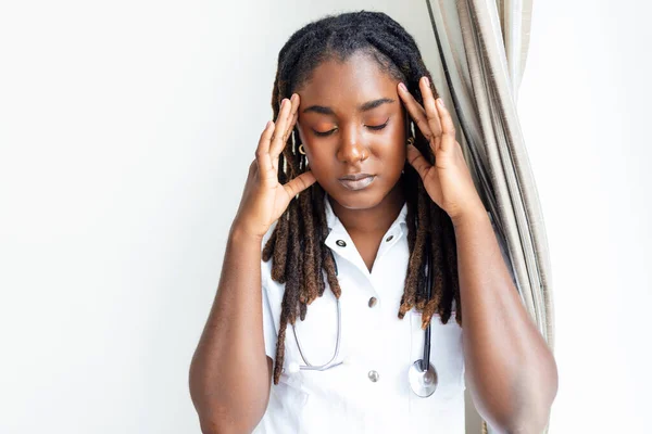 Depressed doctor. Tired doctor near window. Portrait of stressed doctor woman in office. Displeased black healthcare worker. Overworked healthcare worker looking through a window, very concerned, very tired