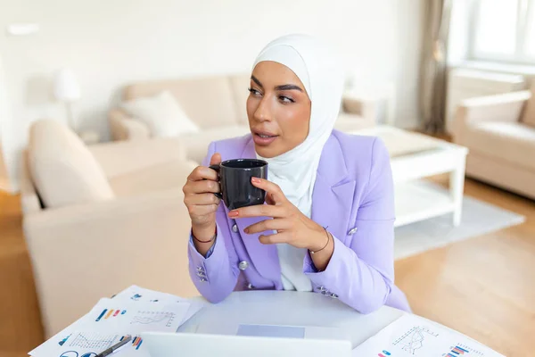 Muslim woman working with computer. Arab Young business woman sitting at her desk at home, working on a laptop computer and drinking coffee or tea. Muslim woman working at a home and using computer.