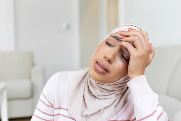 Portrait of an Arabic Muslim woman sitting on a sofa at home with a headache, feeling pain and with an expression of being unwell. Upset woman lying on couch feeling strong headache migraine.