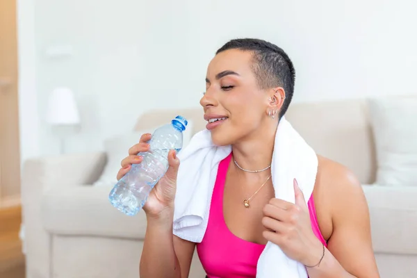 Excited athletic black female in sportswear laughing sitting on floor yoga mat, holding bottle of water with towel on her shoulders