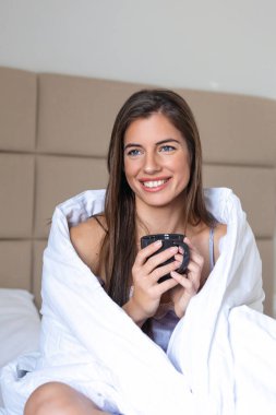 Yatakta uzanırken elinde bir fincan kahve tutan güzel genç bir kadın. Sabah konsepti. Genç bir bayan yatakta dinlenirken ve evde sıcak bir içecek içerken gülümsüyor ve başka tarafa bakıyor.