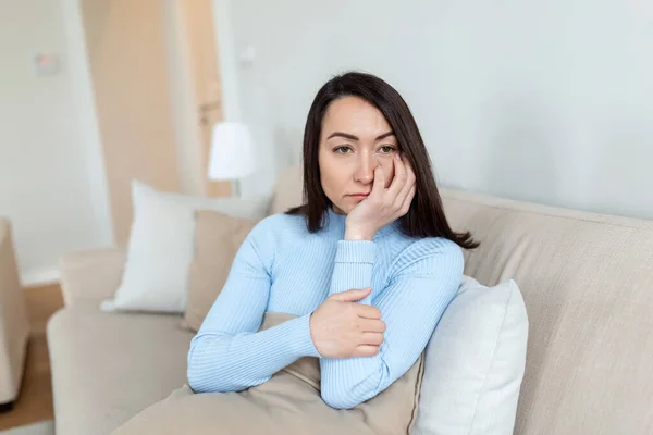 Frustrated Asian lady sitting on sofa, cuddling pillow, looking away at window. Lost in thoughts unhappy stressed woman regretting of wrong decision, spending time alone in living room.