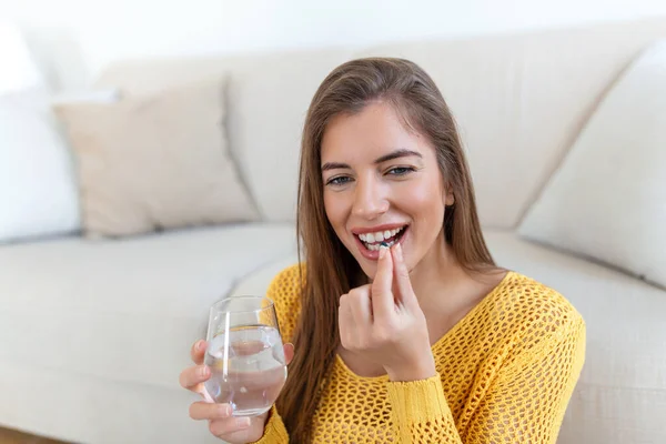 Head shot smiling young woman holding pill and glass of fresh pure water. Healthy millennial lady taking antioxidant medicine vitamins, beauty supplements for hair skin nails, healthcare concept.