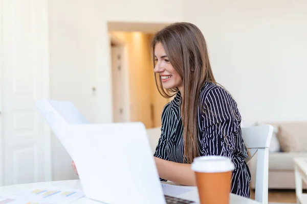 Remote job, technology and people concept - happy smiling young business woman with laptop computer and papers working at home office during the Covid-19 health crisis.