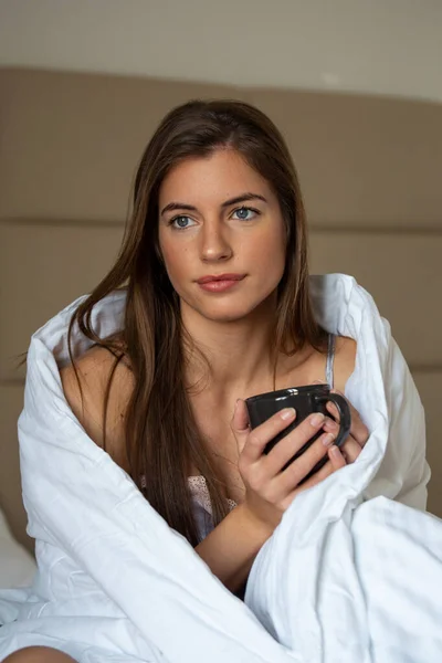 Beautiful woman wrapped in a blanket after waking up, entering a day with cup of coffee happy and relaxed after good night sleep. Sweet dreams, good morning, new day, weekend, holidays concept