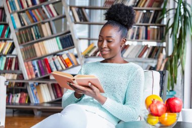 Genç ve güzel bir Afro-Amerikalı kız kanepede kitap okuyor. Arkasında kütüphane rafları var. Güzel bir kadın beyaz bir kanepede kitap okuyor.