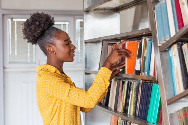Kütüphanede kitap arayan genç siyahi bir kadın. İnsanlar, bilgi, eğitim ve okul konsepti - kütüphanede kitabı olan mutlu öğrenci kız veya genç kadın