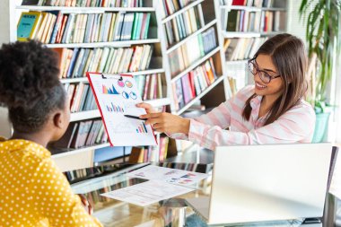 Genç profesyoneller, dizüstü bilgisayar ve tablet kullanarak şirkette çalışıyorlar. Masada iki kadın var, gülücüklerle konuşuyorlar, gümüş ekrana bakıyorlar ve analitiği ellerinde tutuyorlar..