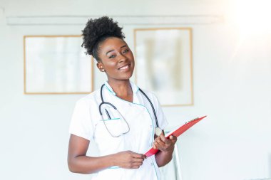 Steteskoplu bir doktor elinde bir defter tutuyor. Hastanede ayakta dikilirken panoda tıbbi form dolduran bir kadın doktorun yakın çekimi.