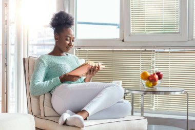 Rahat bir kanepe ve güzel bir kız, kitap okuyor, ev kavramları ve rahatlık, yazı için bir yer. Evdeki pencerenin yanında kitap okuyan güzel siyah bir kadın. Evde kal.