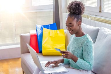 Kanepede laptopla alışveriş yapan genç bir kadın. Genç siyahi kadın online alışveriş yaparken kart bilgilerini giriyor. Çevrimiçi alışveriş kavramı