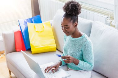 Kadının elinde kredi kartı ve dizüstü bilgisayar var. İnternetten alışveriş konsepti. Genç Afrikalı Amerikalı kız kredi kartıyla moda ürünleri alıyor.