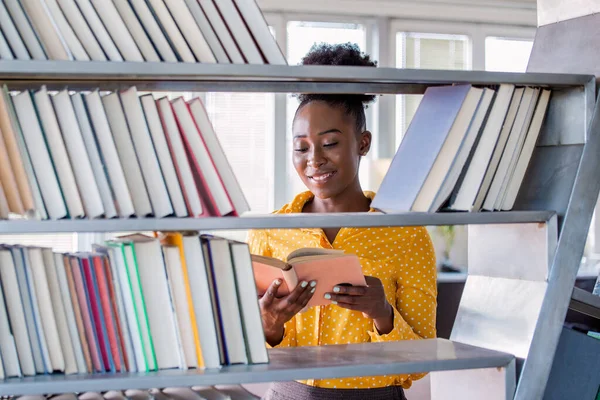 Woman African American college student working at library holding a book in hands, looking smart. bookshelves at the library.