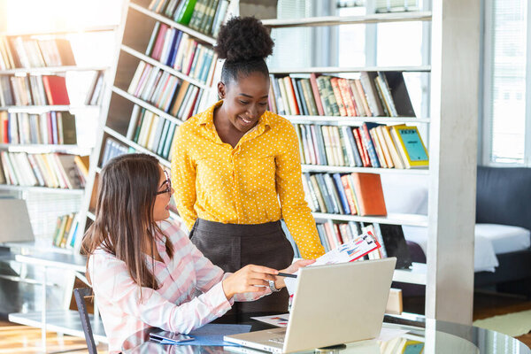 Коллеги по бизнесу работают с ноутбуком, обсуждая бизнес-идеи в современном офисе. Одна девушка сидит за столом, а другая стоит рядом и помогает..