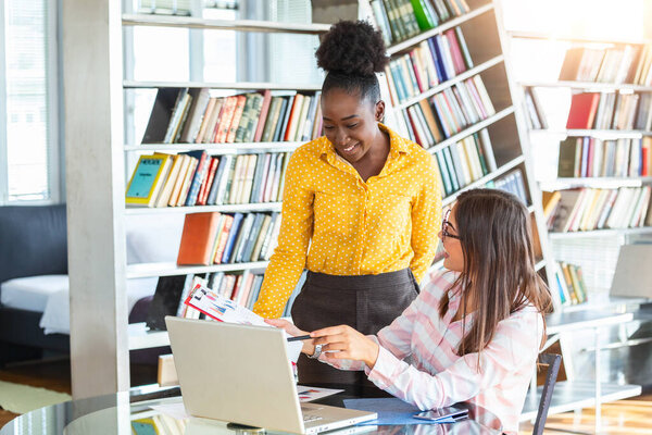 Коллеги по бизнесу работают с ноутбуком, обсуждая бизнес-идеи в современном офисе. Одна девушка сидит за столом, а другая стоит рядом и помогает..