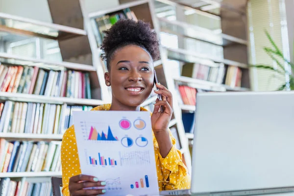 Female businessmen in casual wear work at the desk about accounting and business plan analysis. Young black woman talking on phone at her desk in an office. Graphs and charts