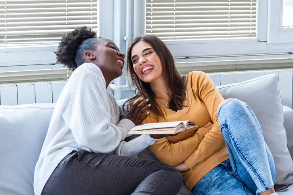 Two women laughing about funny stories from books at home. Friendship and care, happy girl friends. African American and caucasian best friends