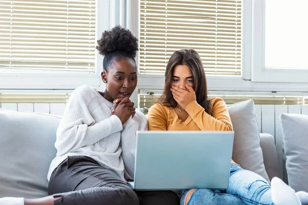 Frightened young women watching movie at home. Girls watching a terror movie on tv sitting on a couch at home. Girlfriends watching scary movie on laptop at home