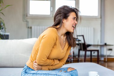 Elleri kasıklarına baskı yapan genç hasta bir kadın. Tıbbi ya da jinekolojik sorunlar, sağlık hizmetleri kavramı. Evdeki kanepede otururken karın ağrısı çeken genç bir kadın.