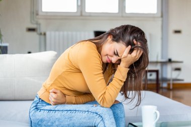 Young sick woman with hands holding pressing her crotch lower abdomen. Medical or gynecological problems, healthcare concept. Young woman suffering from abdominal pain while sitting on sofa at home