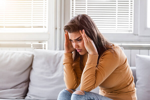 Portrait of a young brunette girl sitting on the couch at home with a headache and migraine . Beautiful woman suffering from chronic daily headaches. Sad woman holding her head because sinus pain