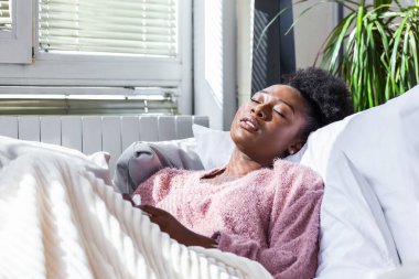 Sick young african woman feeling cold covered with blanket sit on bed, ill black girl shivering freezing warming at home wrapped with plaid, no central heating problem, fever temperature flu concept