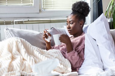 Hastalık, mevsimsel virüs problemi konsepti. Kanepede yatıp termometredeki sıcaklığa bakarak hasta olan Afrikalı Amerikalı kadın. Hasta kadın yüksek ateşle yatakta yatıyor. Soğuk algınlığı ve migren.