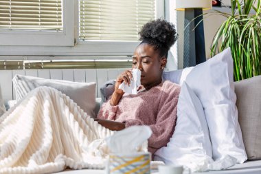 Hasta kadın. Flu. kadın üşüttü. Dokuların içine hapşırmak. Baş ağrısı. Virüs. İlaçlar. Burnunu mendile sümküren soğuk algınlığına yakalanmış genç bir kadın. Kanepede oturan, başı ağrıyan hasta kadın.