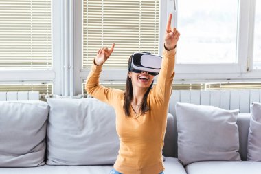 Sanal gerçeklik gözlüklü kadın. Evde kanepede VR kulaklık takan gülümseyen genç bir kadın. Kadın evde sanal gerçekliğin tadını çıkarıyor. Sanal gerçeklik kulaklığı takan Arayüzlü Güzel Kadın.