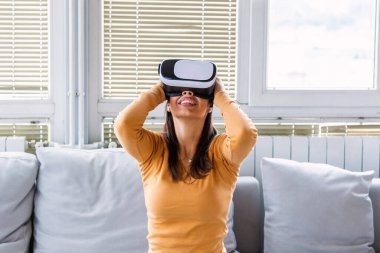 Sanal gerçeklik gözlüklü kadın. Evde kanepede VR kulaklık takan gülümseyen genç bir kadın. Kadın evde sanal gerçekliğin tadını çıkarıyor. Sanal gerçeklik kulaklığı takan Arayüzlü Güzel Kadın.