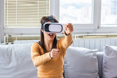Kanepeye oturup sanal gerçeklik gözlüğü takan, internet sitesine bakan ve el hareketli 3 boyutlu simülasyon ekranı kullanan zarif bir kadın. Yeni gerçekliğin tadını çıkarıyorum. VR kulaklıklı çekici genç kadın.