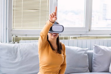 Sanal gerçeklik gözlüklü kadın. Evde kanepede VR kulaklık takan gülümseyen genç bir kadın. Kadın evde sanal gerçekliğin tadını çıkarıyor. Sanal gerçeklik kulaklığı takan Arayüzlü Güzel Kadın.