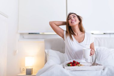 Pijamalı çekici bir kadın sabah kalktığında kahve ve taze kahvaltıyı yatakta, ahşap tepside yapıyor.