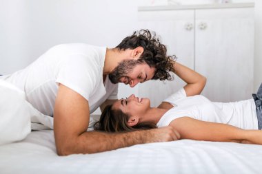 Sevecen genç ve çekici bir çift evdeki yatak odasında romantik bir anı paylaşıyor. Mutlu genç çift evde yatak odasında yatarken sarılıyor ve gülümsüyor..