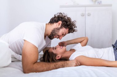 Sevecen genç ve çekici bir çift evdeki yatak odasında romantik bir anı paylaşıyor. Mutlu genç çift evde yatak odasında yatarken sarılıyor ve gülümsüyor..