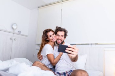Sabah selfie 'si. Genç mutlu çift yatakta akıllı telefon ve kucaklaşmayla kendi portresini yapıyor, Yatakta Tatlı Fotoğraflar Çeken Çift