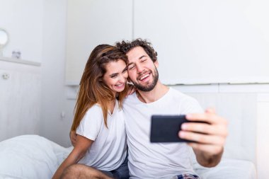 Yatakta Selfie çeken genç çift cep telefonuyla yatakta güzel ve mutlu bir çift akıllı telefonla kendi portresini yapıyor ve sarılıyorlar.,