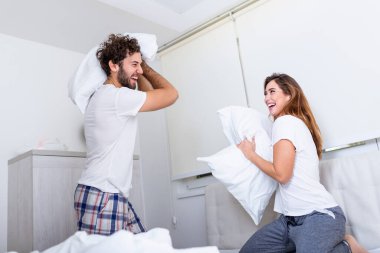 Çift yastık savaşı yaparken eğleniyor. Genç mutlu çift evdeki yatak odasında yastıkları döver..