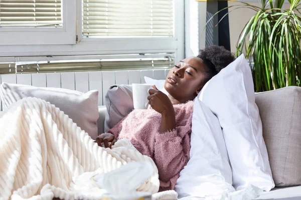 Sick African American woman with headache sitting under the blanket and drinking hot beverage. Sick woman covered with a blanket lying in bed with seasonal infections, flu, allergy resting.