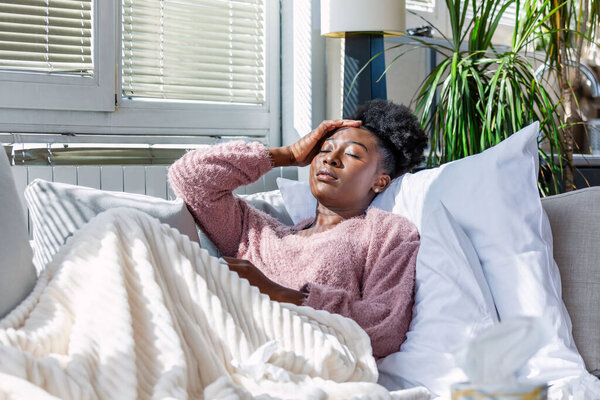 Больной афроамериканка с головной болью сидит под одеялом. Больная женщина, покрытая одеялом, лежит в постели с сезонными инфекциями, гриппом, аллергия отдыхает.