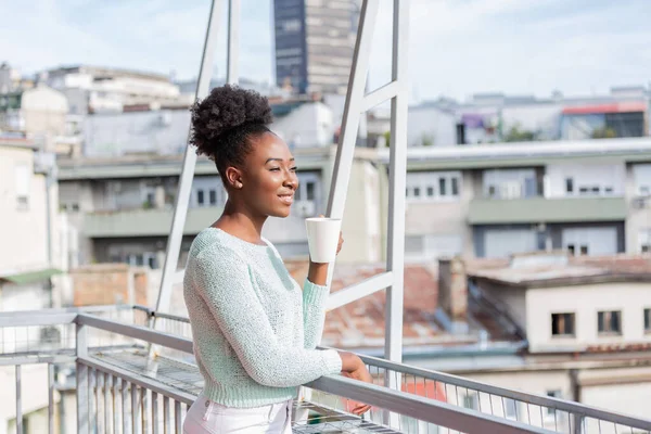 Portrait of a happy african american black woman thinking and looking away with coffee on vacation with a resort or hotel with city view in the background