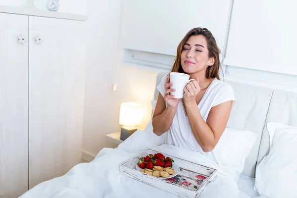 Breakfast in bed for young beautiful woman. Woman having coffee and breakfast in bed with fruits, coffee and biscuits. Morning waking up in hotel
