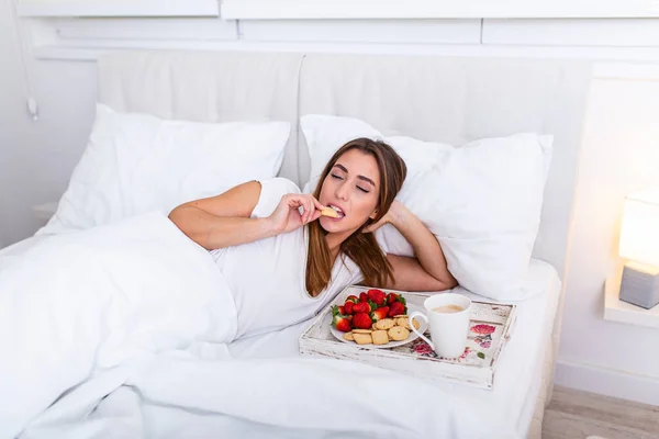 Breakfast in bed for young beautiful woman. Woman having breakfast in bed with fruits, coffee and biscuits. Morning waking up in hotel