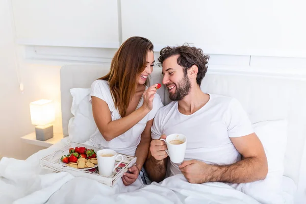 Cute couple having breakfast in bed in the bedroom. Beautiful woman feeding her boyfriend strawberries in bed while having breakfast and coffee in bedroom