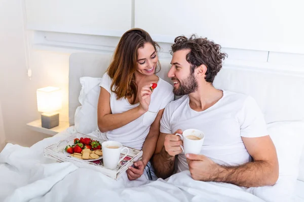 Cute couple having breakfast in bed in the bedroom. Beautiful woman feeding her boyfriend strawberries in bed while having breakfast and coffee in bedroom