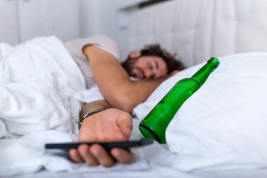 Sarhoşken mesajlaşmak. Genç adam, elinde boş içki şişesiyle körkütük sarhoş halde yatakta yatıyor. Alkolle sarhoş olmuş. Alkolizm alışkanlık sarhoşluk tehlikeli alışkanlık konsepti